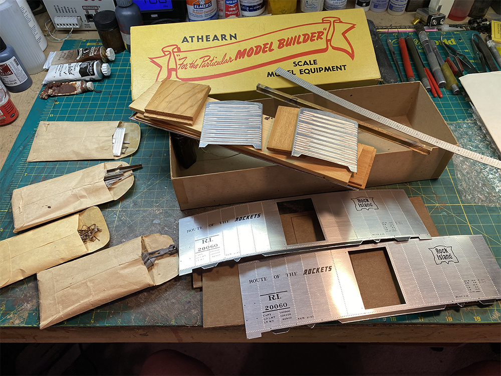 Workbench with green top covered with small tan envelopes, wood and metal toy train boxcar parts, and yellow box lid with red and black printing.