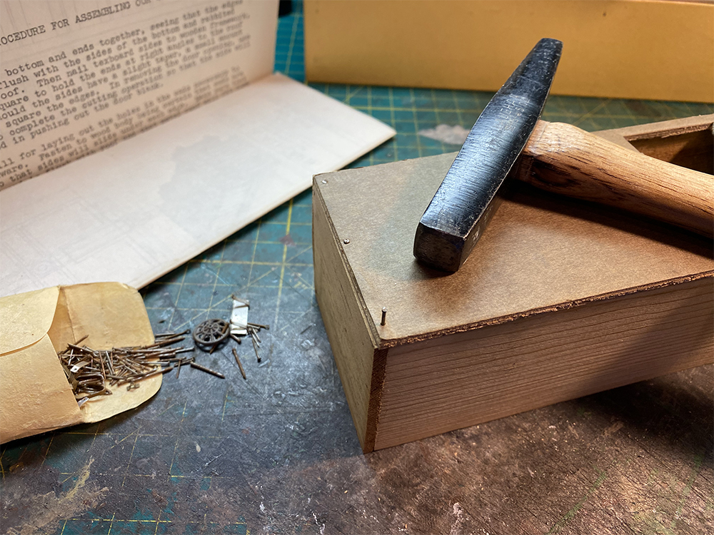 Build an O scale freight car: Small tan envelope containing small nails and other metal parts on green surface next to a brown wood box. The box has a small black hammer resting on top of it.