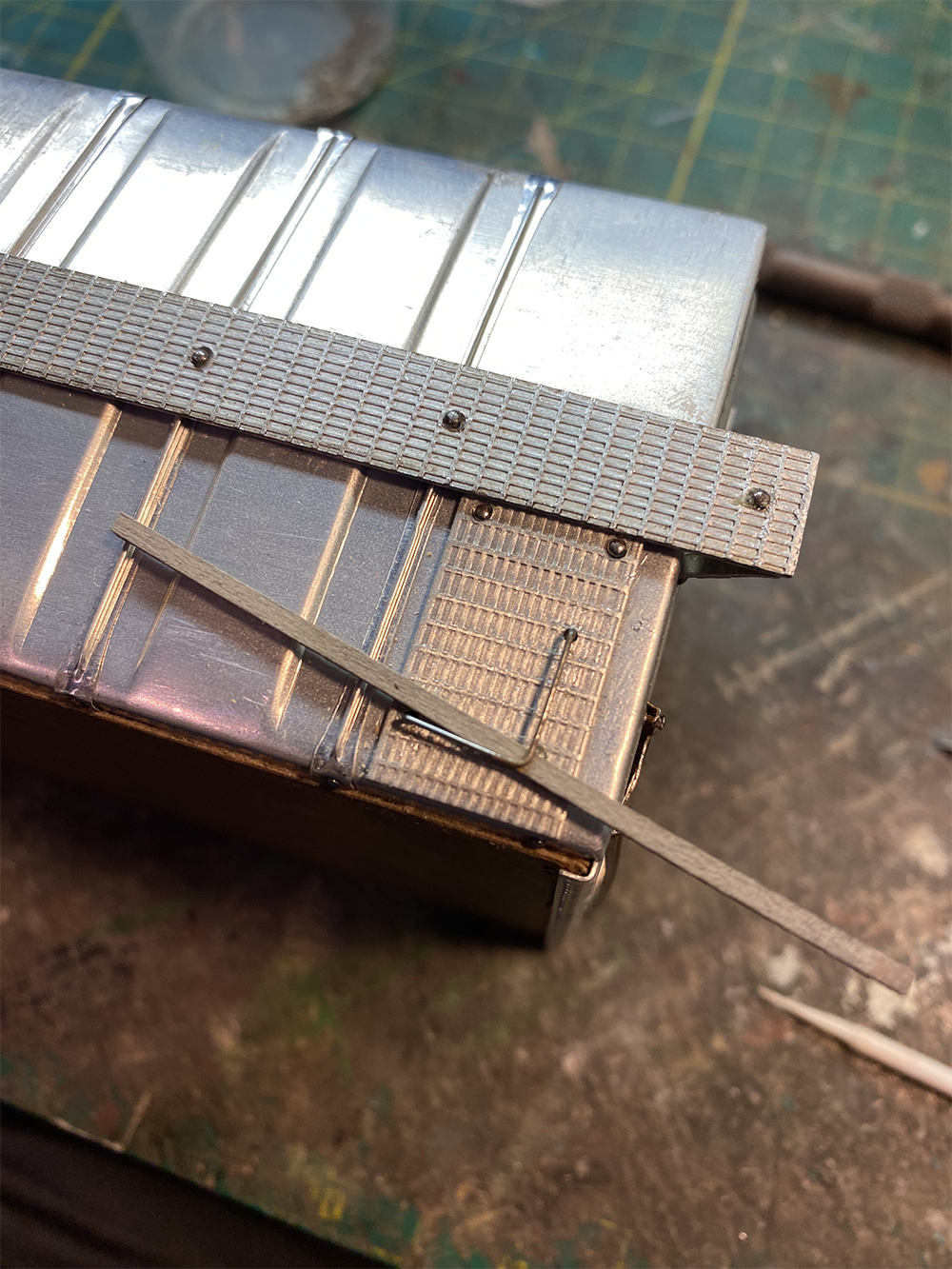 Build an O scale freight car: Silver corrugated metal with metal stamped running board detail and sliver wire bent in L shape supported by small piece of wood.