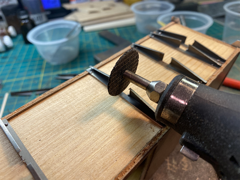 Build an O scale freight car: Black motor tool with silver collet and brown cutting wheel suspended over notch in triangular metal frame section attached to brown wood floor of model train car.