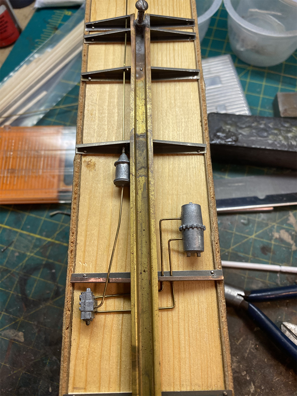 Build an O scale freight car: Wood model train boxcar floor with silver and brass metal frame sections and braces, as well as bare brass wire and three gray metal rectangular castings.