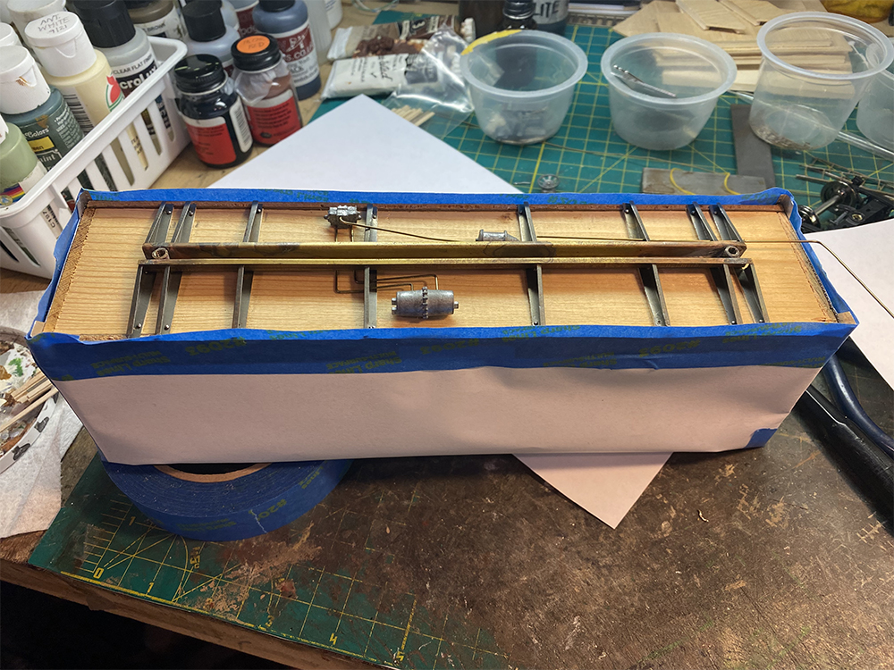 Wood and metal model train boxcar floor with silver and brass pieces surrounded by blue tape and white paper resting on a crowded workbench containing all manner of bottles tools and small parts in clear-plastic containers.