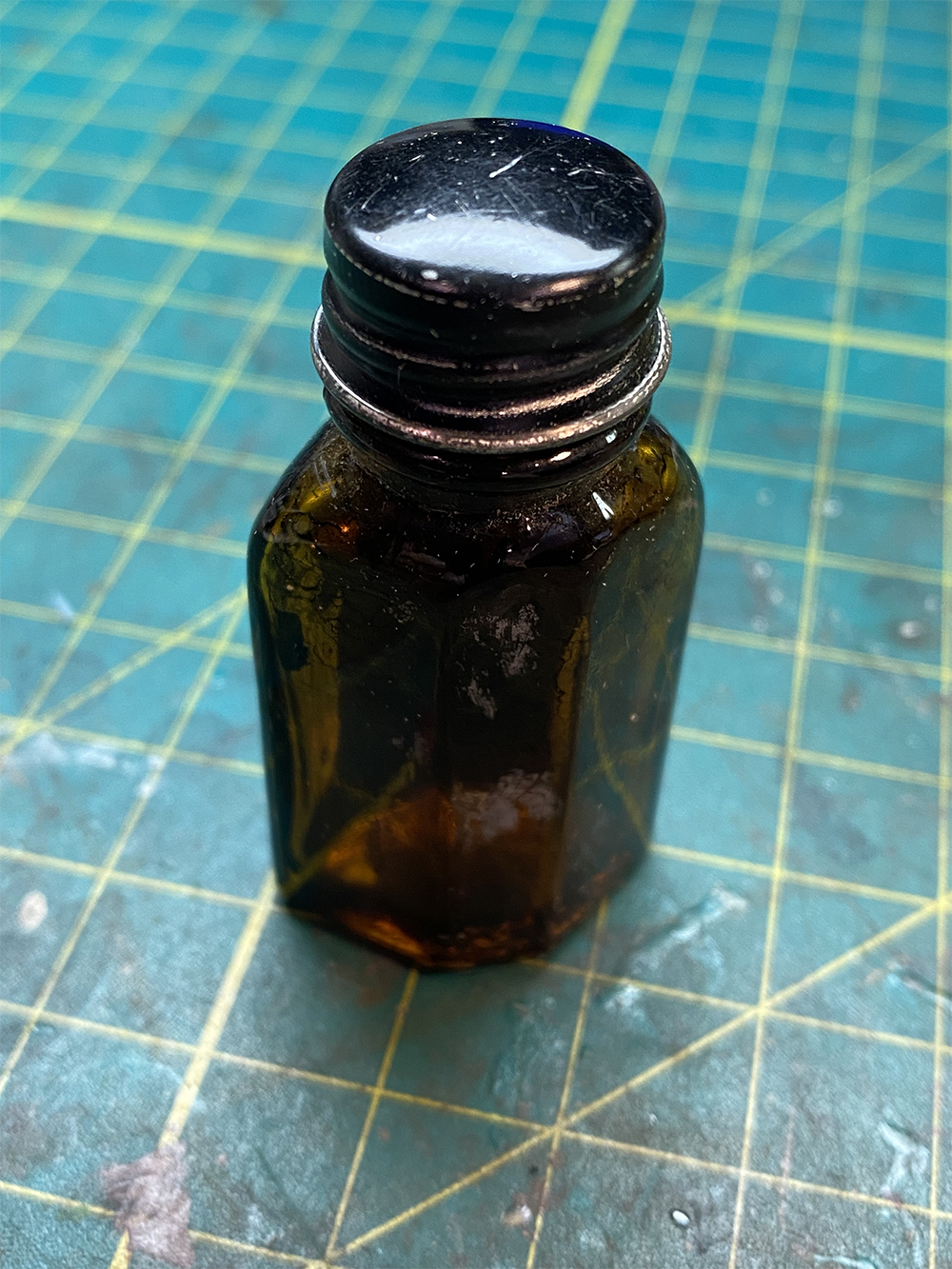 small brown glass bottle with black metal cap resting on a green and yellow checkered workbench mat.