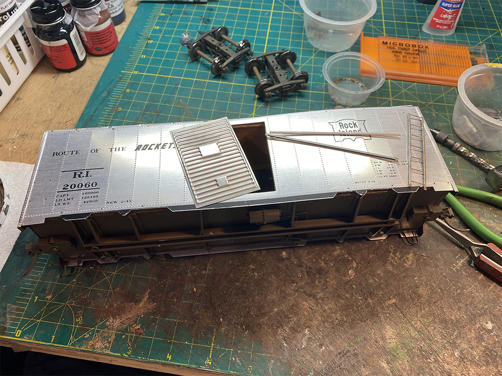 Silver metal toy train boxcar sides with corrugated metal door and formed metal door tracks resting on green workbench top.
