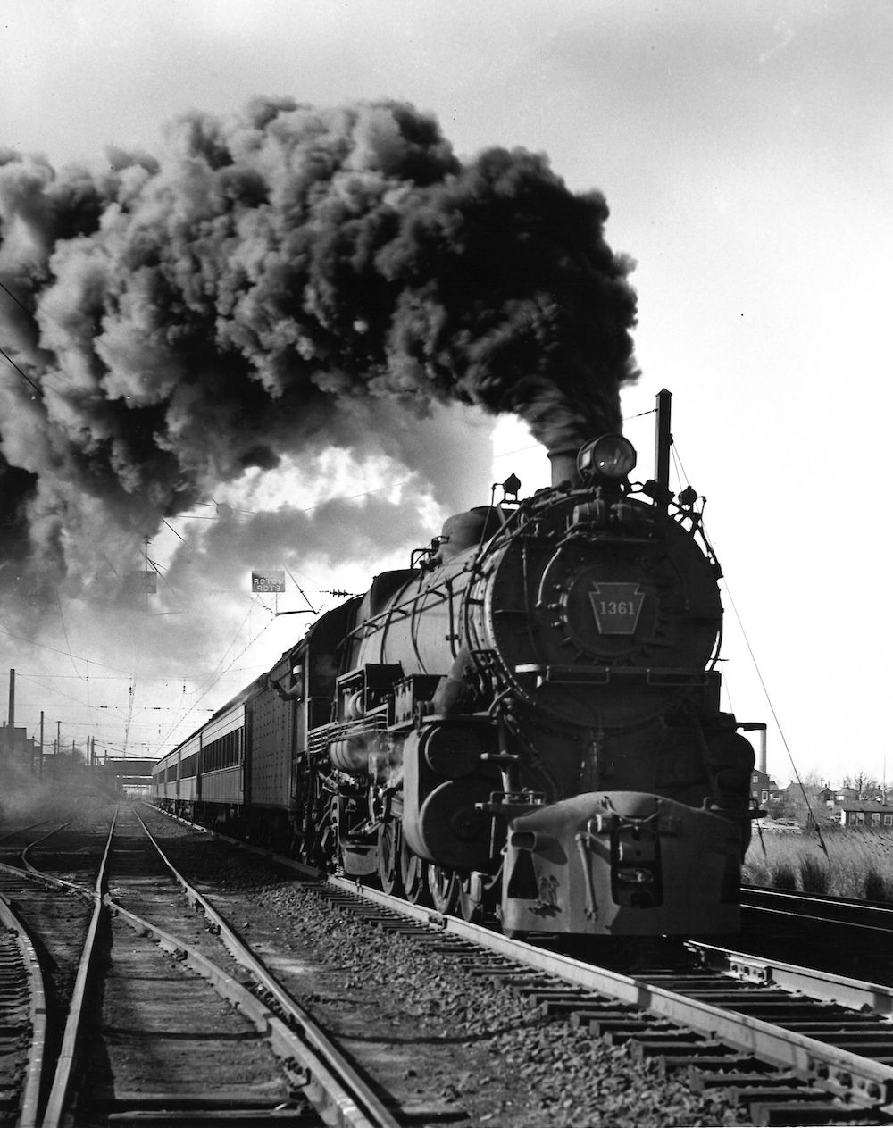 Black and white photo of steam-powered passenger train departing town.