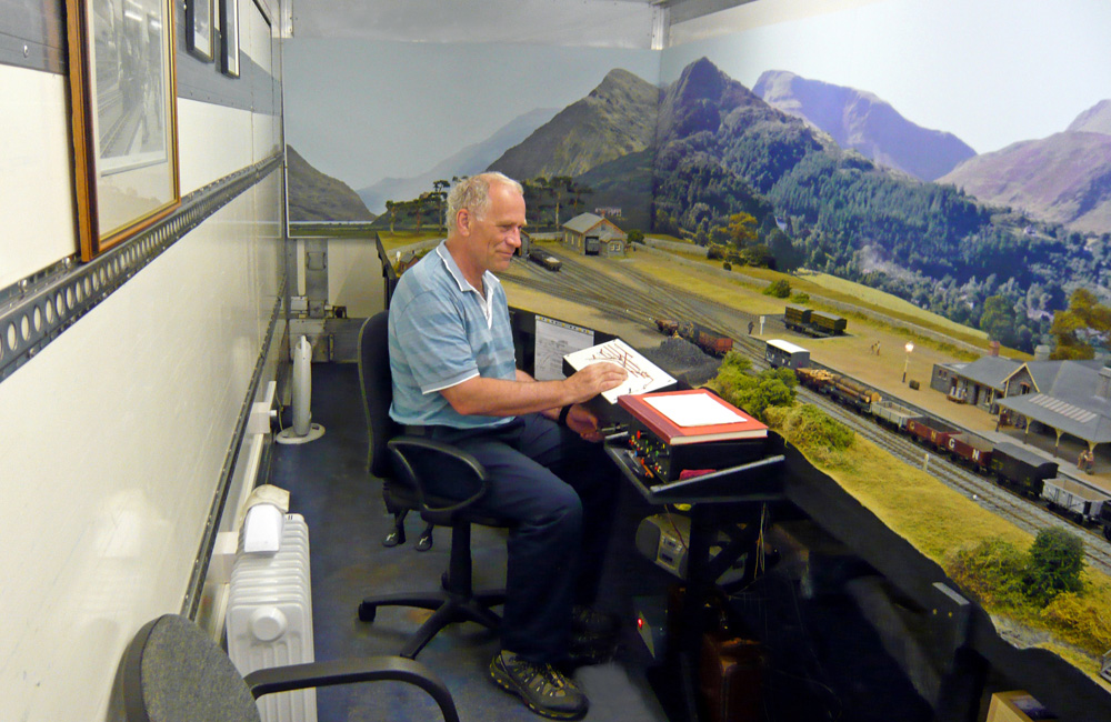 A seated man operates a model railroad in a long, narrow space
