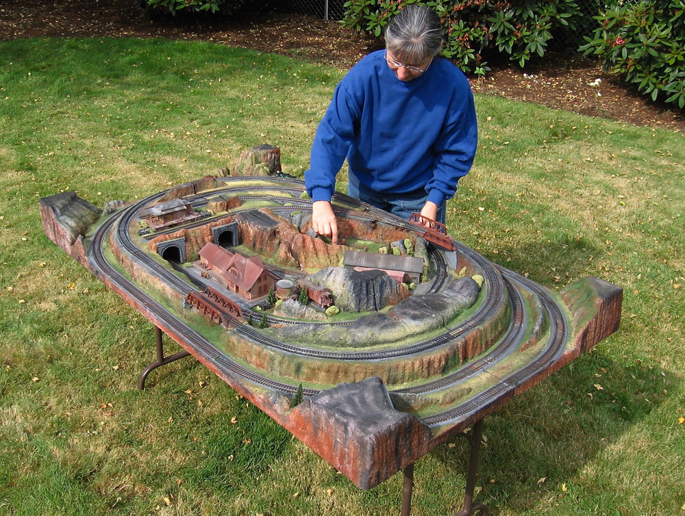 A man sits next to a nearly finished train layout outside.