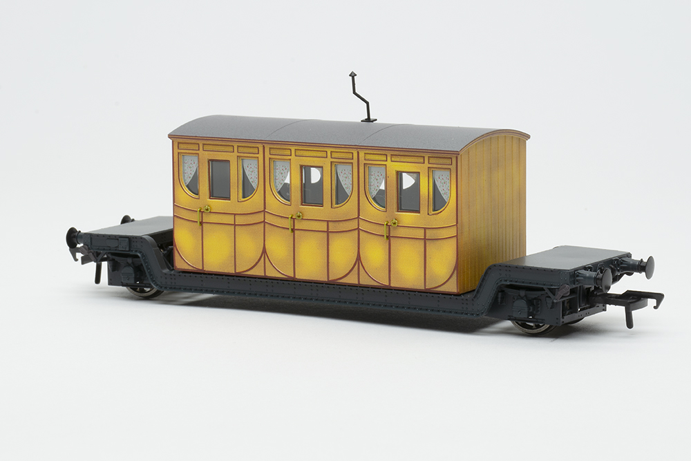 Rapido UK Titfield Thunderbolt: Black model flat car with two wheels and yellow railway coach body mounted on top.