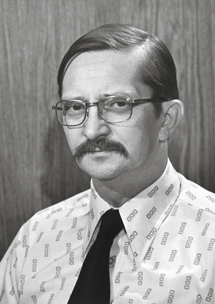 Black-and-white photo of man with moustache and glasses wearing a dress shirt and necktie.