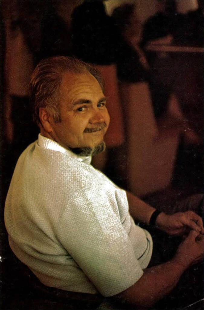 Color image of man with gray hair and round beard smiling while looking over shoulder.