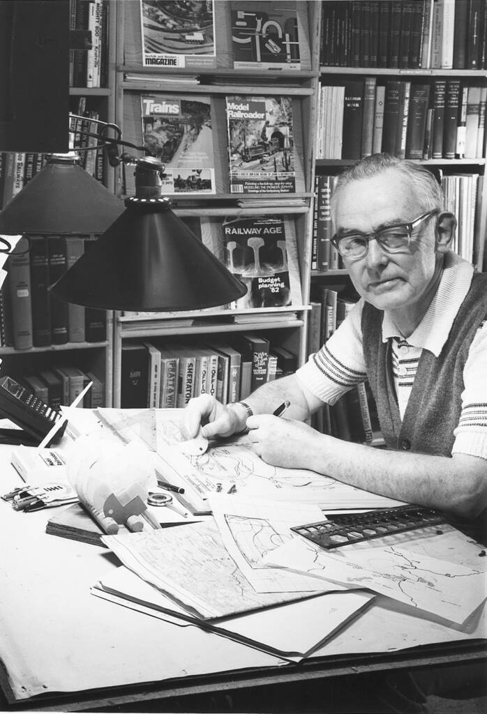 Black-and-white photo of man at drafting table with books and magazines in background.