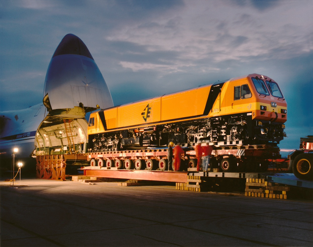 An orange European-design diesel locomotive mounted on a multi-wheeled road trailer, is loading into a giant cargo aircraft