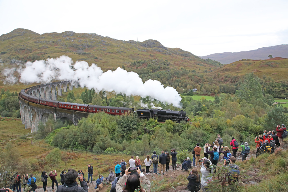 Steam train on curved viaduct with large number of people taking photos in foreground