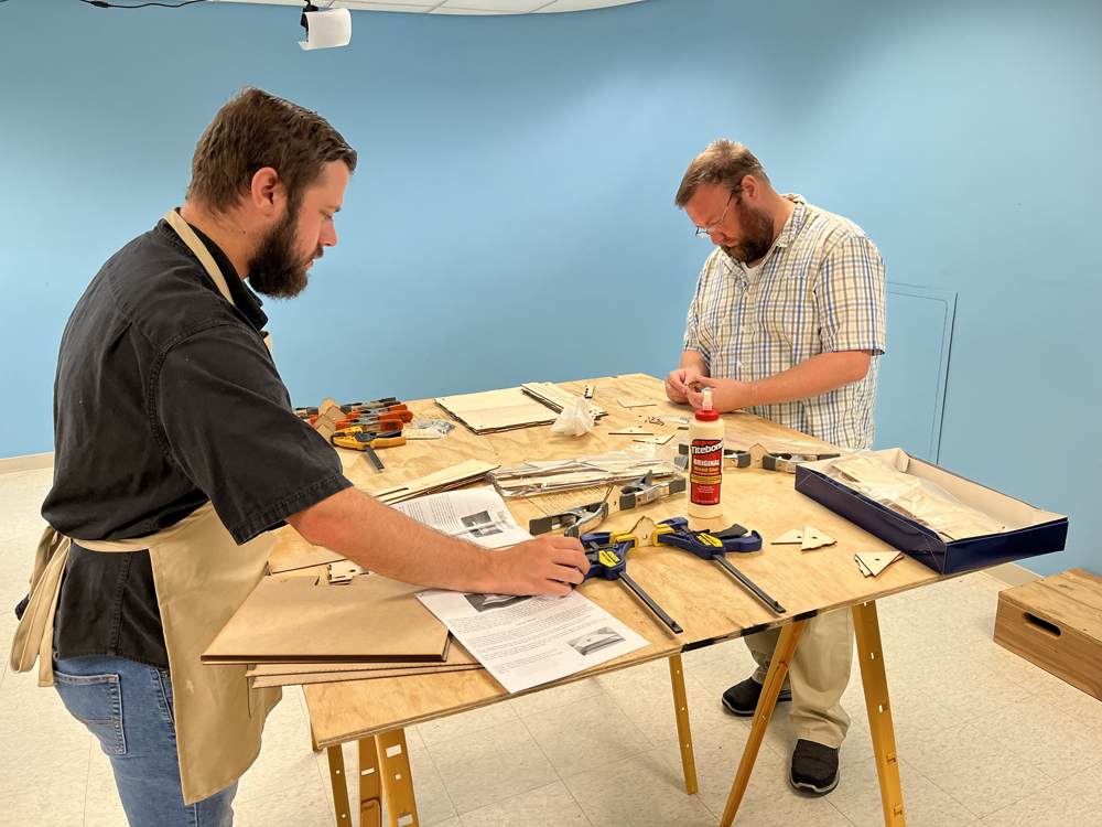 Two men with beards working at make-shift workbench set up on yellow sawhorses using assorted hand tools and clamps to build small brown wood boxes.