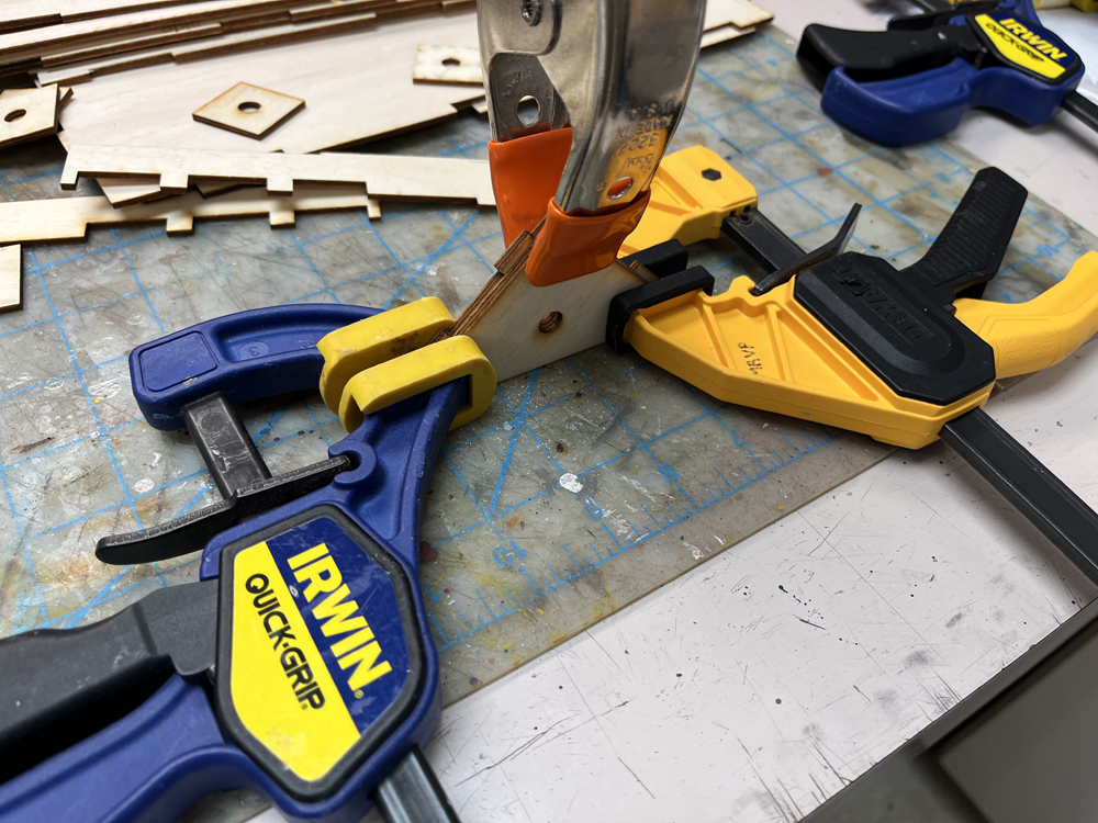 Yellow, blue, black, and orange clamps holding wood triangle on gray workbench mat.