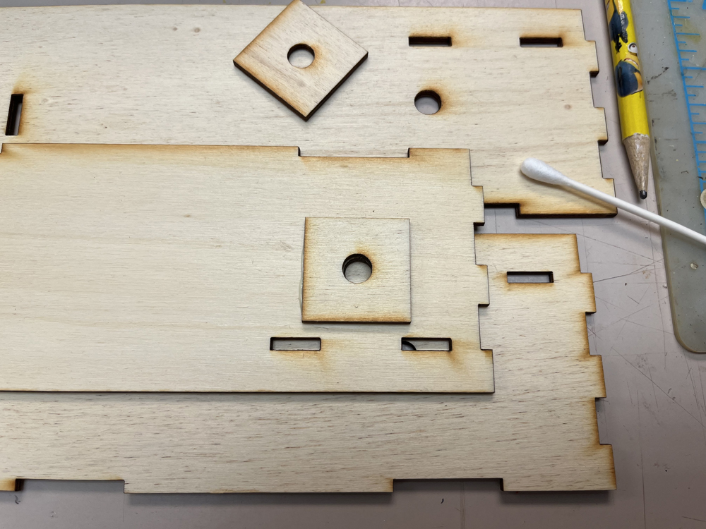 Wood squares with center holes resting on wood rectangular parts on gray workbench, along with white cotton swab and yellow pencil.