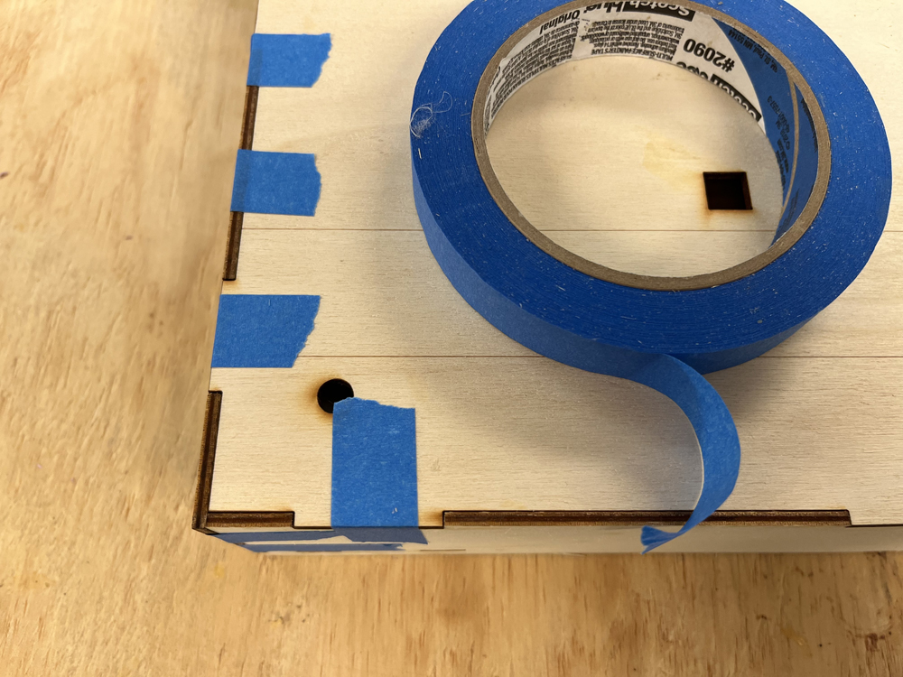 Roll of blue tape on wood box with blue tape pieces holding brown box together.