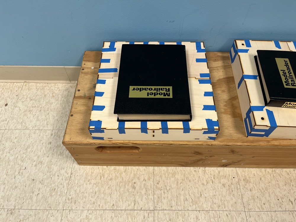 Brown box with blue tape on edges sitting on bench on tan floor. A large, black book sits on top of the brown box.