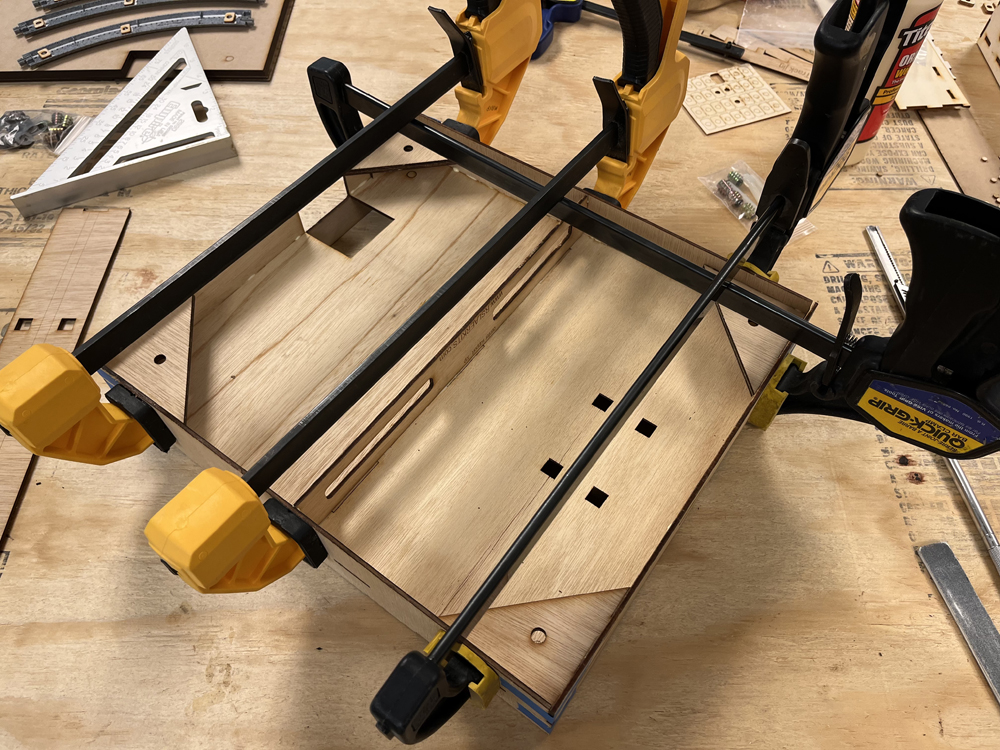 Yellow and black bar clamps holding sides and ends of a wooden box together on a wood workbench top.