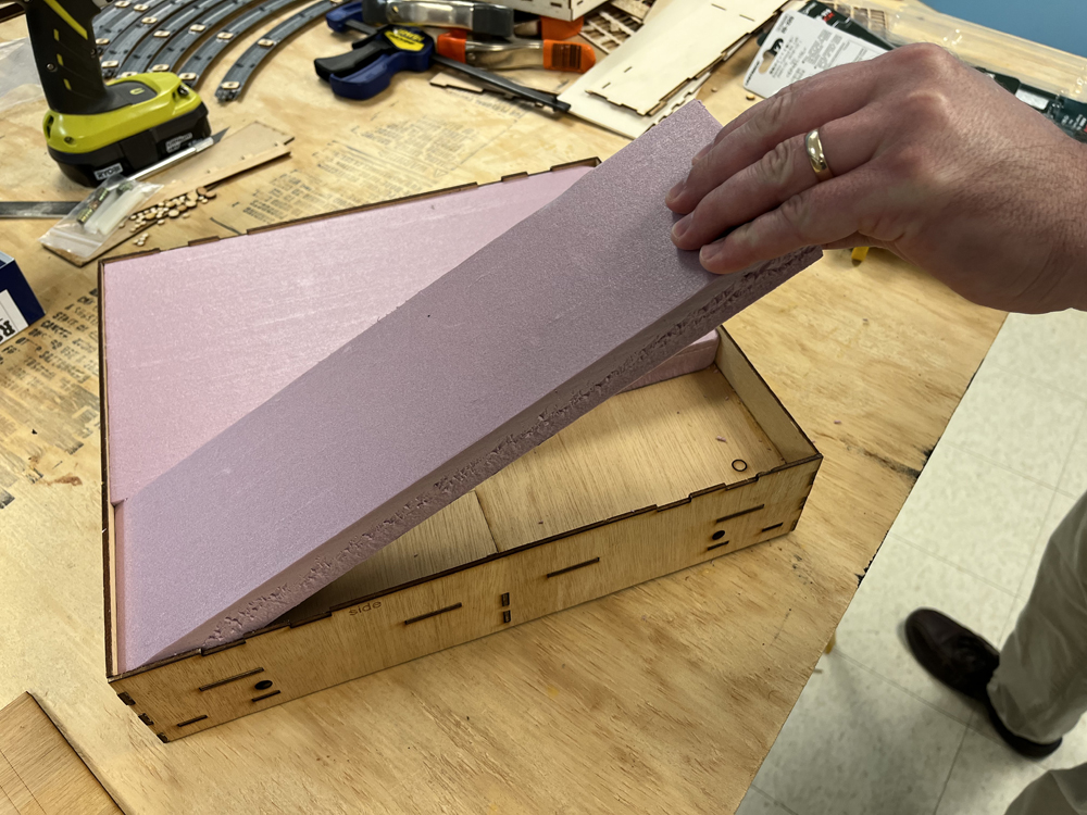 Tan hand wearing gold ring placing pink insulation foam into brown, wooden tray.