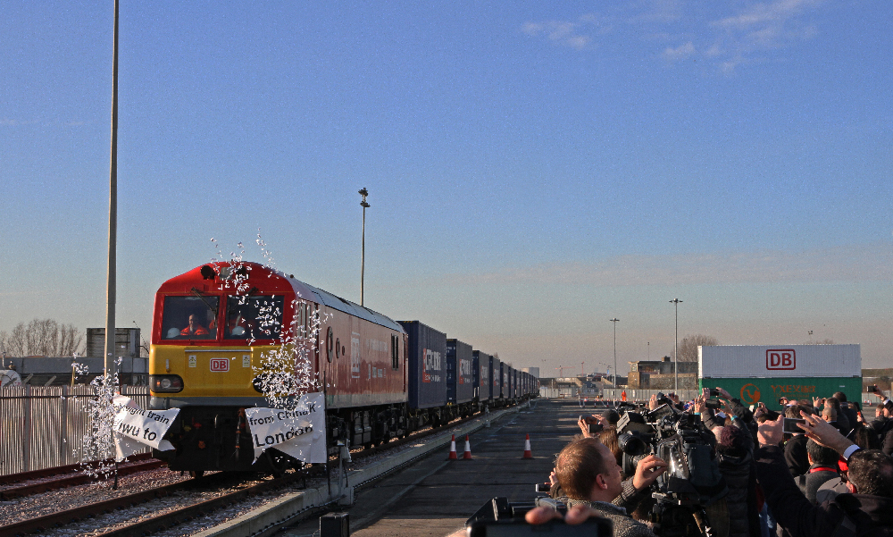 Red European diesel freight locomotive with a container train passing a crowd of people.