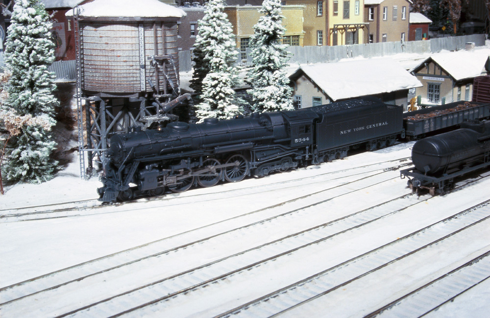 A steam locomotive stands by a station in a snow-covered rail yard