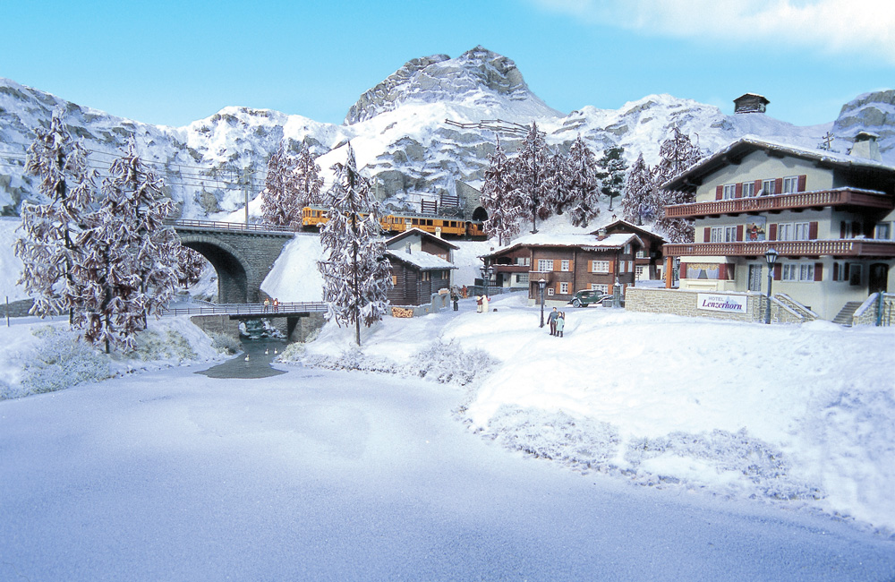A snowy scene with a chalet, a railroad bridge over a frozen river, and distant mountains