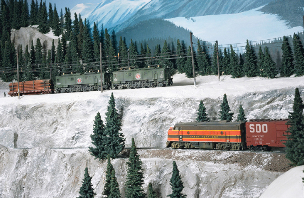 A diesel rolls along a snowy cliff face while an electric locomotive passes on the track above
