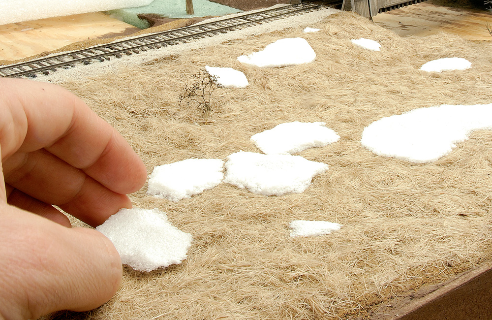 A hand places a white foam snowdrift on a slope of brown grass