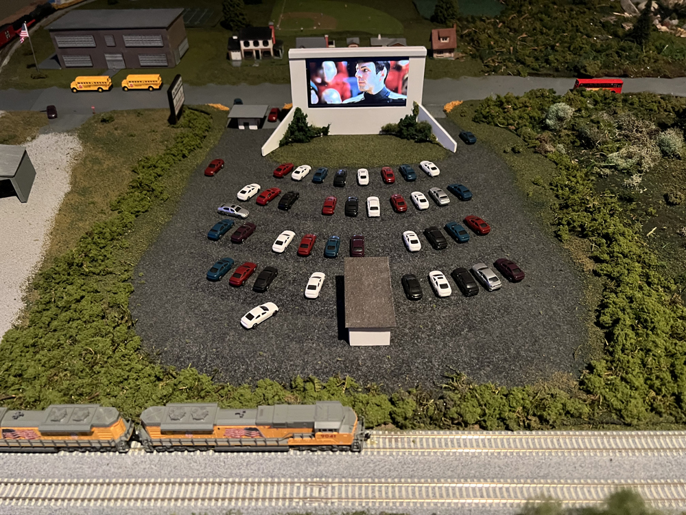 An N scale train passes by behind a drive-in theater