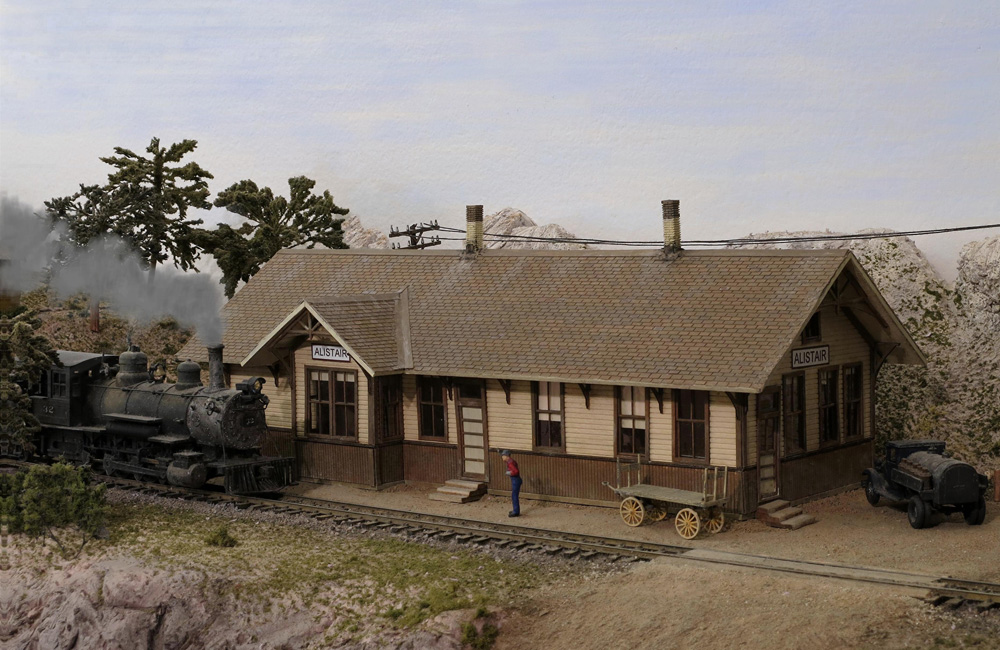 A black steam locomotive approaches a tan-and-brown model wood depot