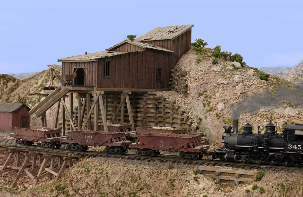 A steam locomotive pushes three wood-sided hopper cars over a trestle in front of a hillside ore mine