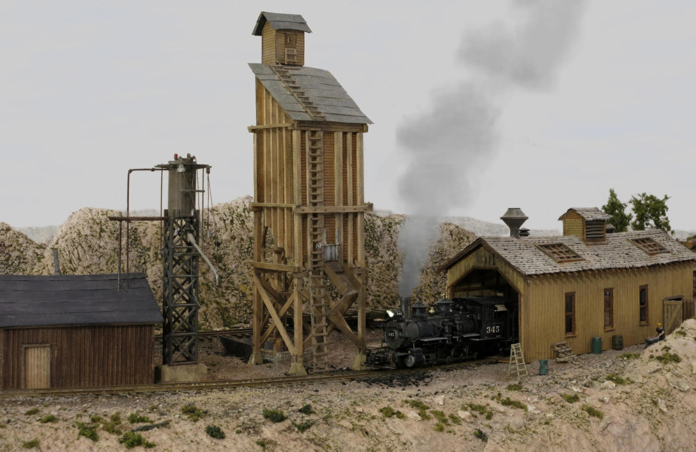 A black steam locomotive rolls out of a wood engine house past a coal tower and a sand house