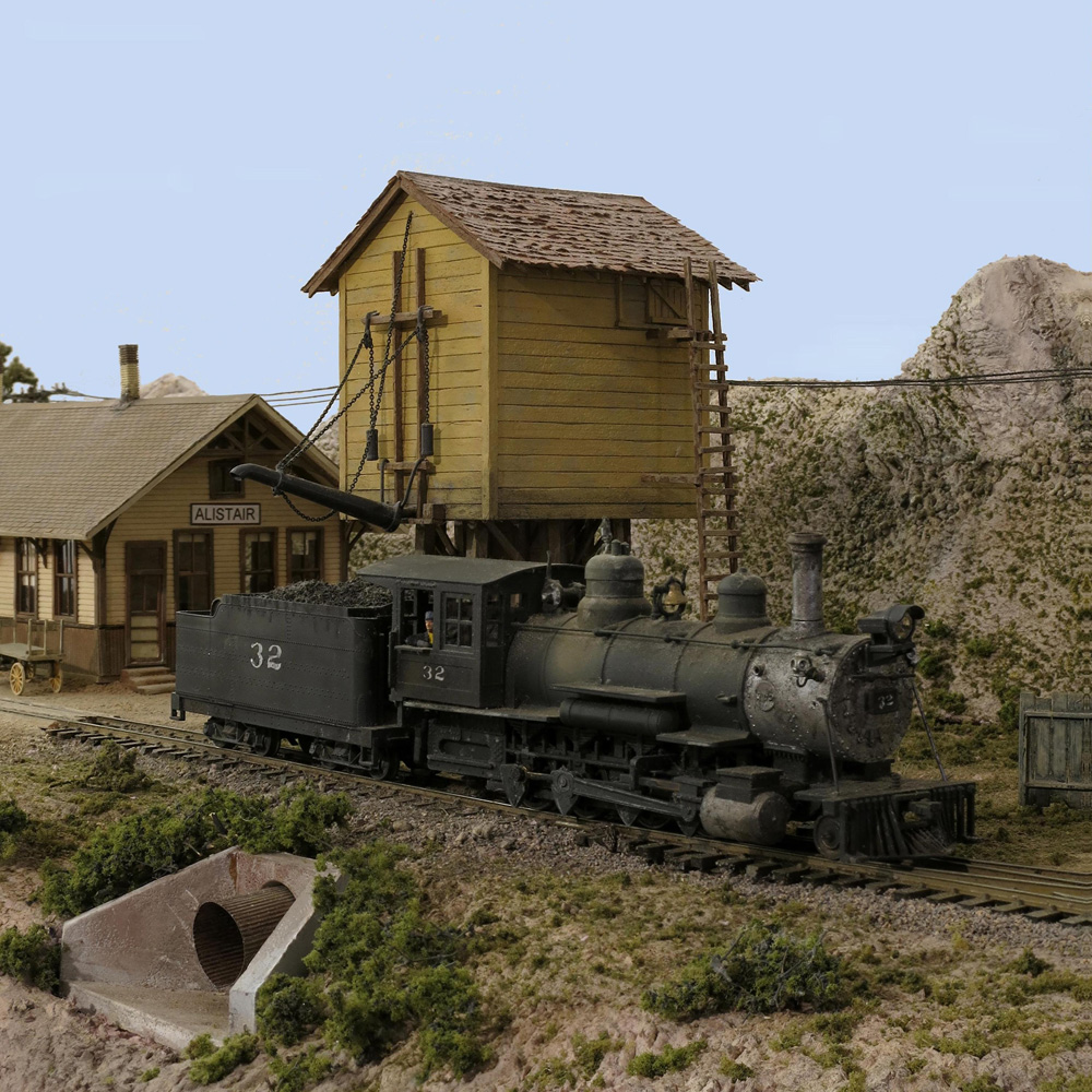A steam locomotive pulls to a stop next to a square yellow wood-sided water tower