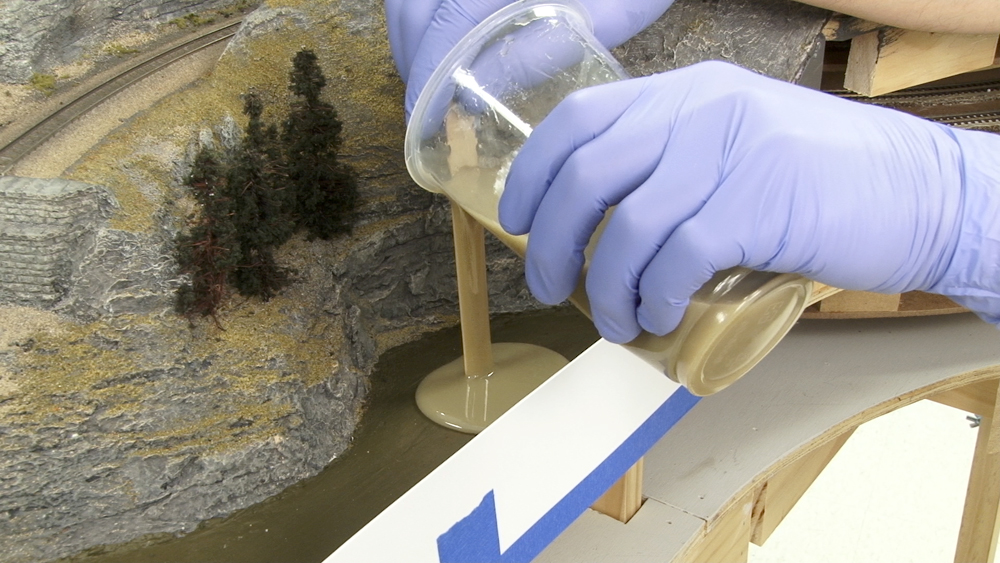 Photo showing gloved hands holding cup with stick guiding resin onto a layout surface.