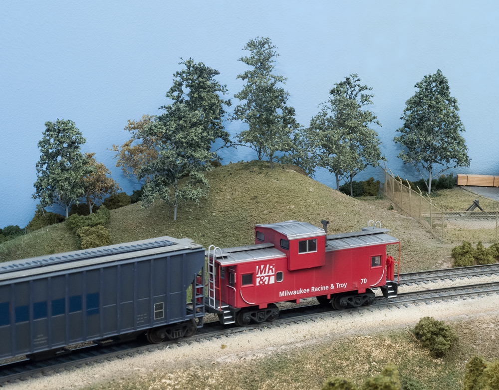 A red caboose and blue covered hopper pass a tree-covered hill on an HO scale layout.