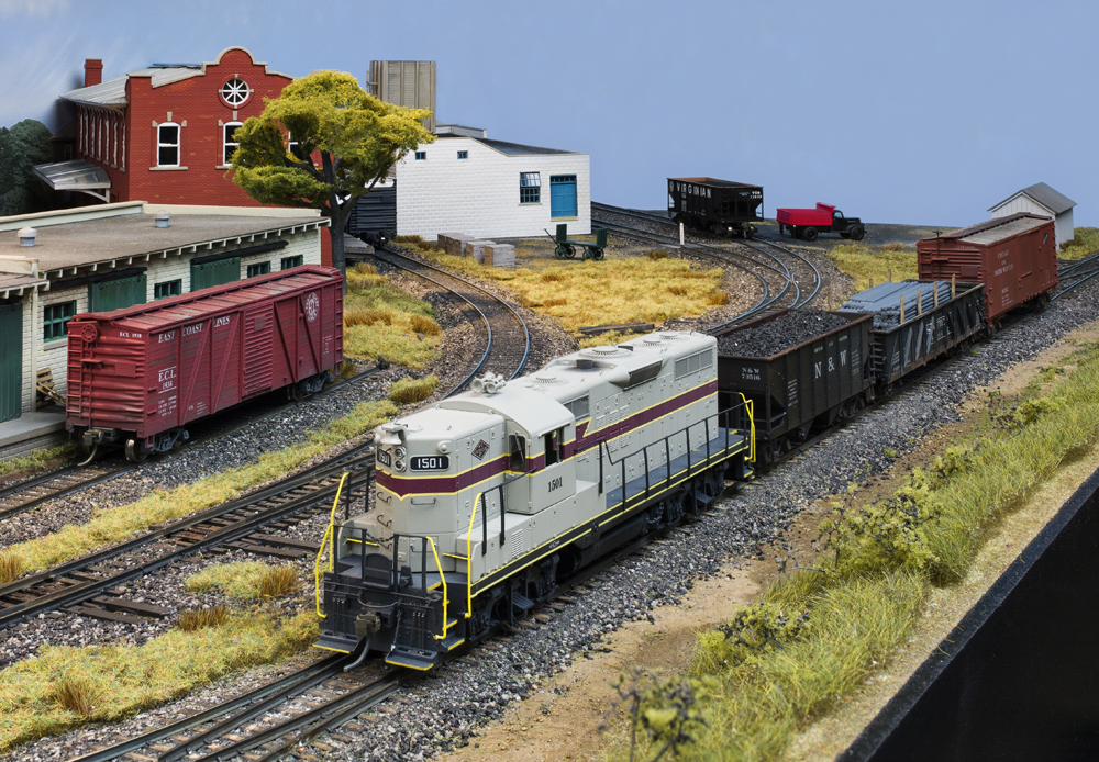Color photo of gray locomotive, freight cars, and structures on HO scale layout.
