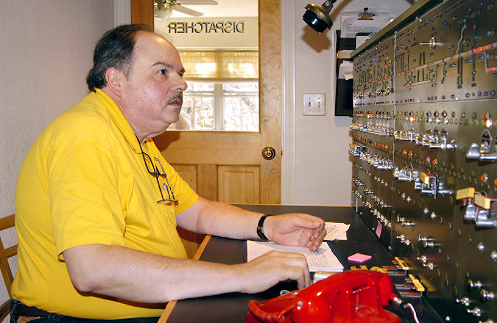 A man sits in front of a control board with switches and lights on it