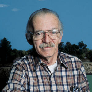 A man in a plaid shirt smiles at the camera in front of a model train layout