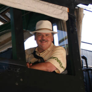 A man in a Panama hat smiles from the cab window of a steam locomotive