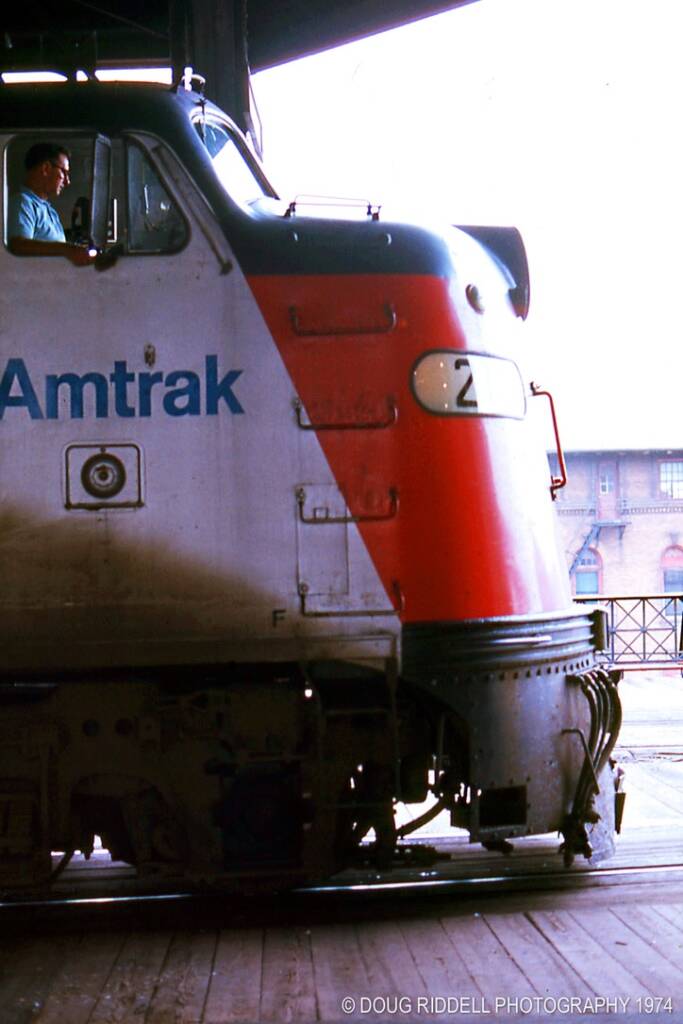 A locomotive engineer in a teal-colored shirt is seen in the cab of a silver-painted Amtrak locomotive standng at a train station platform