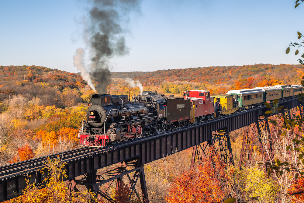Steam powered passenger train crosses trestle bridge with fall foliage in the background.