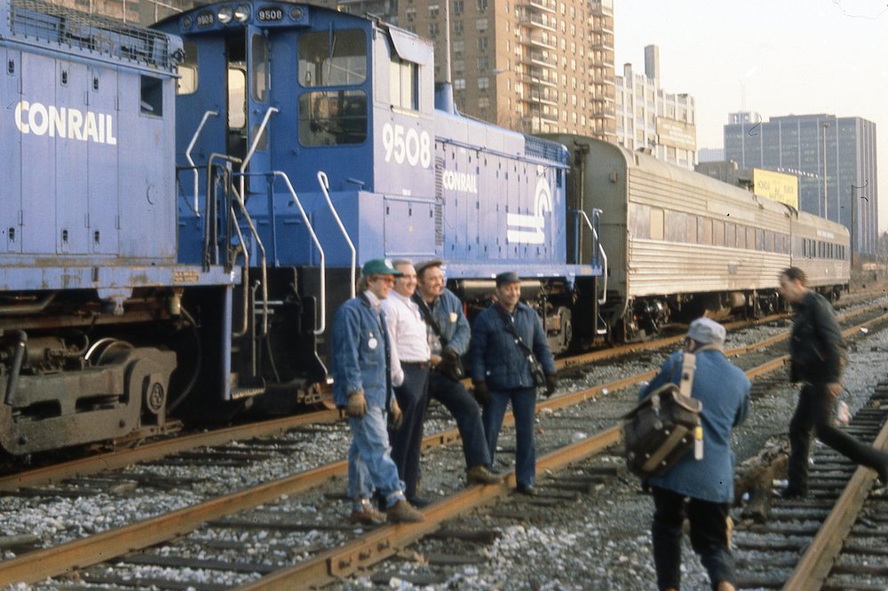 Railroad crew members pose with a diesel engine. Two passenger cars are to the right.