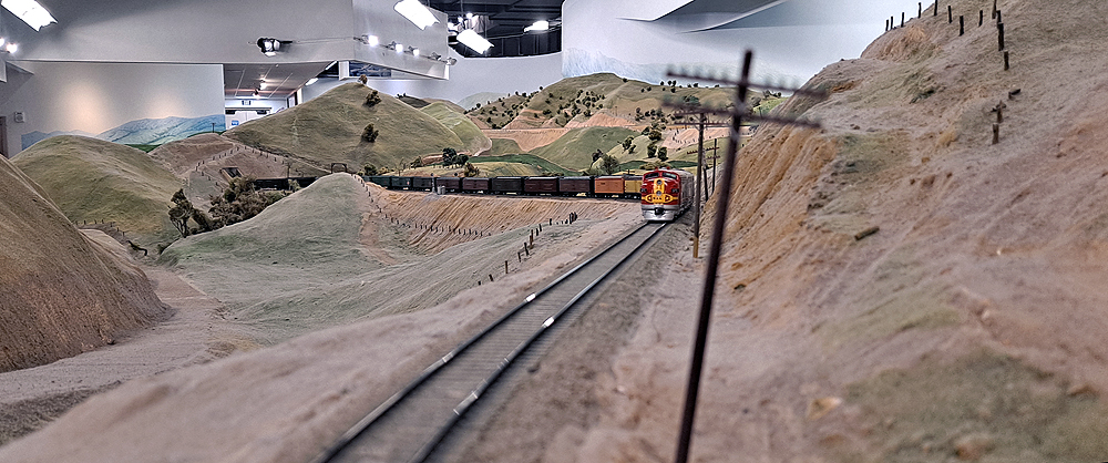 A freight train led by Santa Fe EMD F units in Warbonnet paint climbs a single track through high desert scenery on a large model railroad.