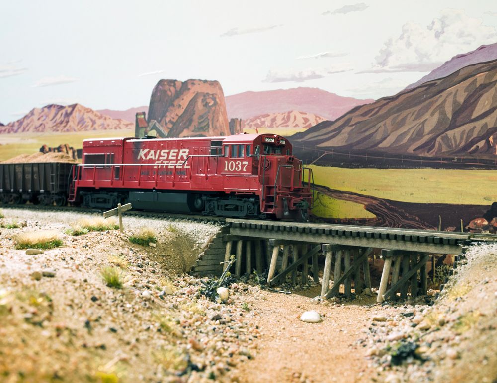 Photo of red HO scale locomotive crossing wood bridge with painted backdrop in background.