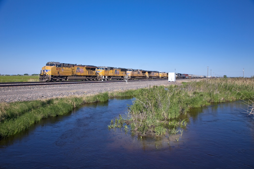 Blue skies are reflected in a water-filled ditch while a yellow train passes in the background