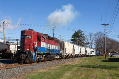 Red, white, and blue high-nose diesel locomotive pulling a train of covered hoppers.