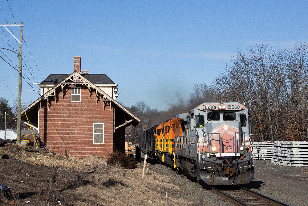 Grey diesel locomotive leads freight train past an old station building.