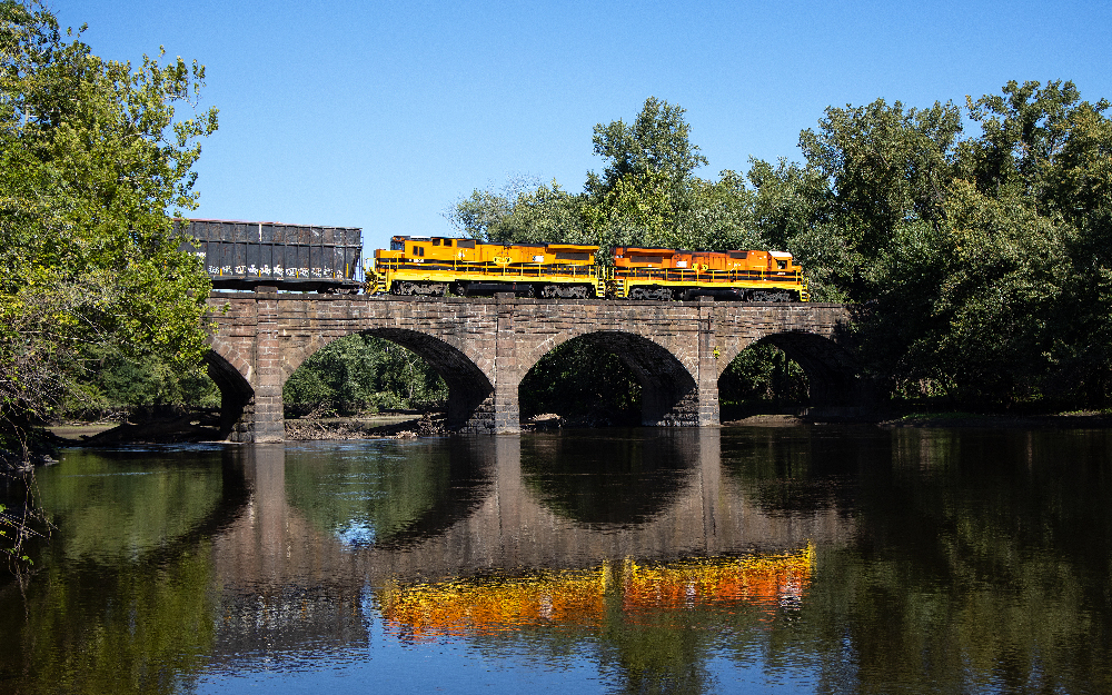Two orange and yellow diesel locomotives pull a freight train across a stone arch bridge.