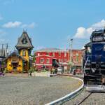 A dark blue diesel locomotive next to a yellow depot with brick buildings beyond